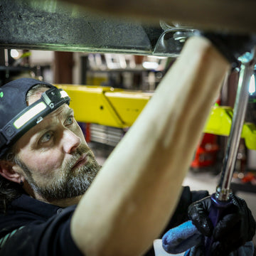 Mechanic using a socket wrench under a vehicle with focused expression and headlamp.