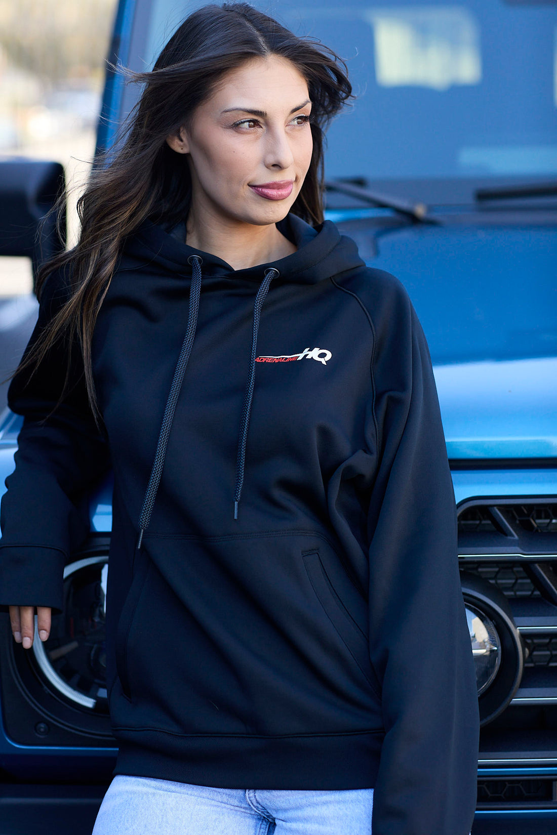 Woman wearing a black hoodie with a small embroidered logo, standing in front of a blue vehicle.