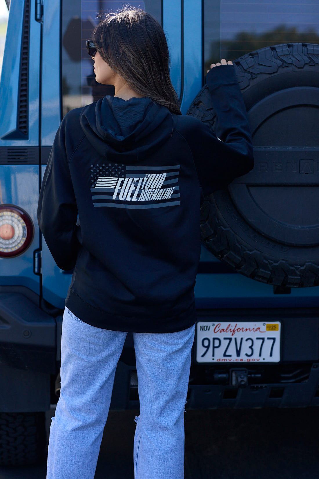 Woman wearing a black hoodie with a small embroidered logo, standing in front of a blue vehicle.