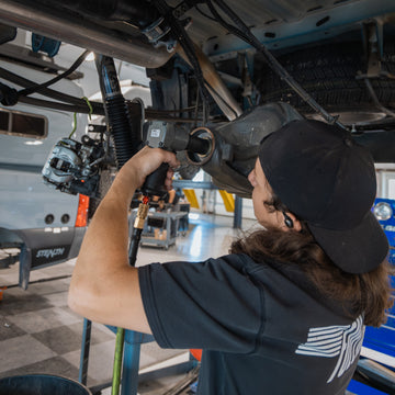 Mechanic using pneumatic tool to install part under a lifted vehicle in a workshop.