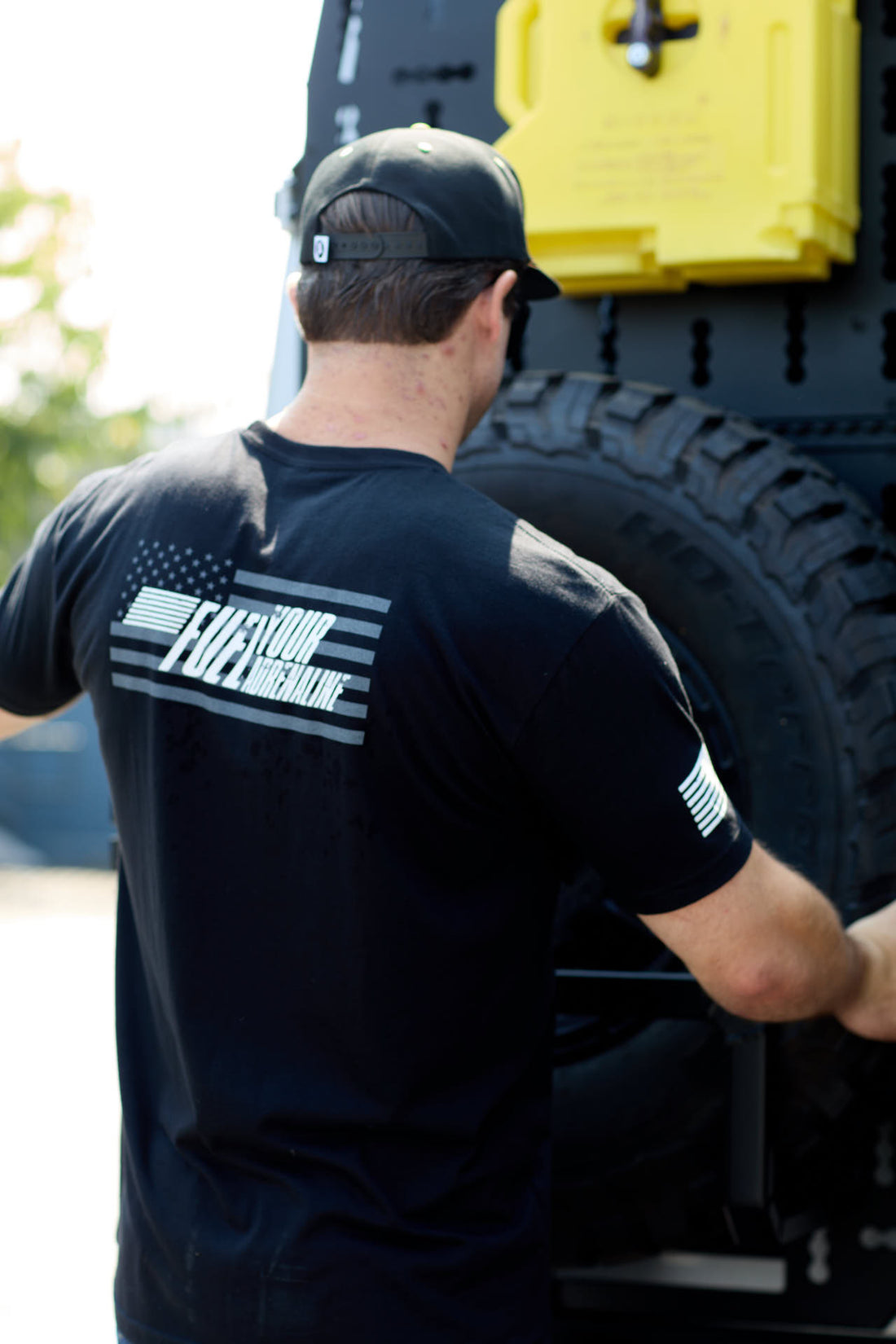 Man outdoors wearing black tee with American flag and 'Fuel Your Greatness' on the back
