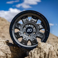 Matte black off-road wheel propped on rocky terrain under a blue sky.