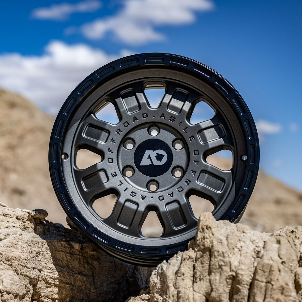 Matte black off-road wheel propped on rocky terrain under a blue sky.