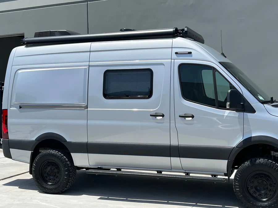 Silver camper van with black off-road tires and roof-mounted awning parked near a gray building.