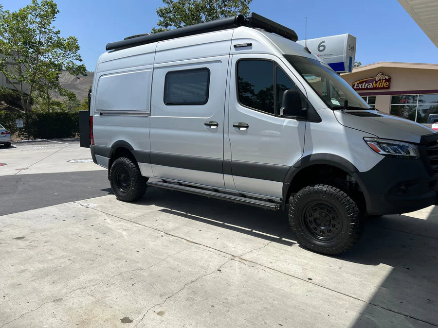 Silver camper van with off-road tires parked beside a gas station on a sunny day.