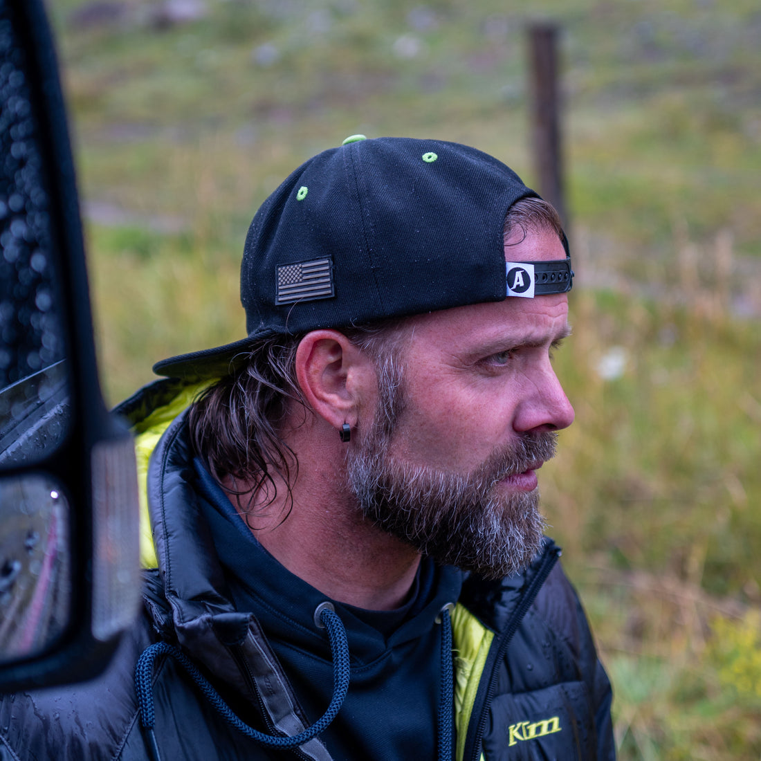 Man wearing a black snapback hat with an American flag patch, standing outdoors in the rain.