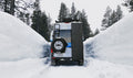 Off-road camper van driving through deep snow between tall snowbanks in a forested area.
