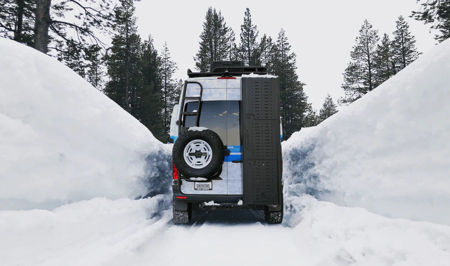 Off-road camper van driving through deep snow between tall snowbanks in a forested area.