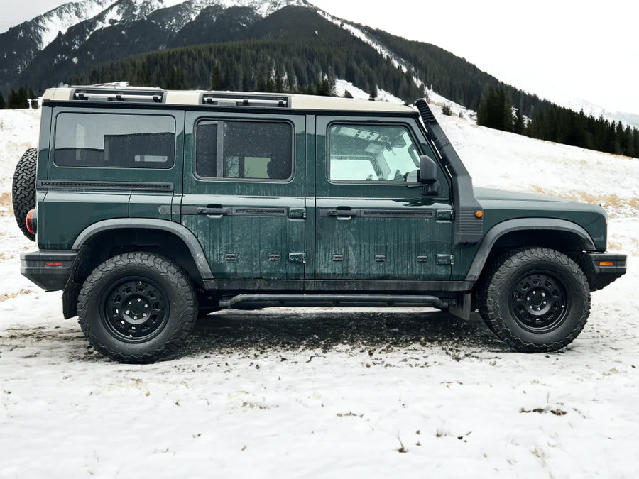 Green off-road SUV parked on snowy mountain terrain with pine trees in background