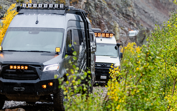 Black and white adventure vans with LED lights driving through a rocky, forested trail.