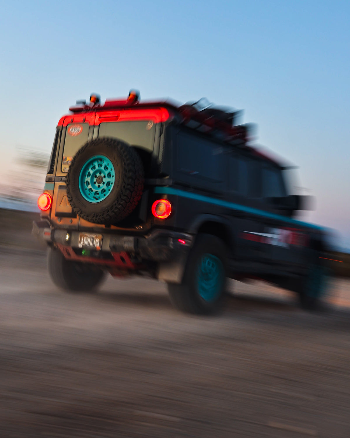 Off-road SUV with roof rack and teal wheels driving on a dirt road at sunset