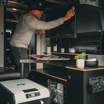 Man cleaning a TV screen inside a modern camper van kitchen setup