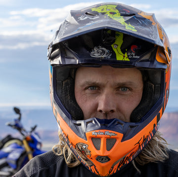 Man wearing an orange and black motocross helmet with a motorcycle in the background