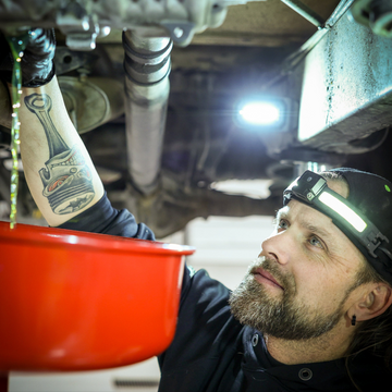 Man wearing headlamp draining oil from vehicle into red container in garage setting
